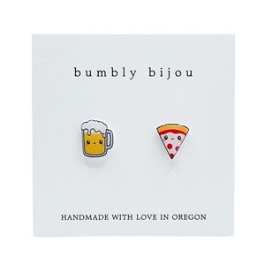 May include: A pair of earrings featuring a cartoon beer mug and a pizza slice. The earrings are handmade with love in Oregon and are sold by the shop "bumbly bijou".