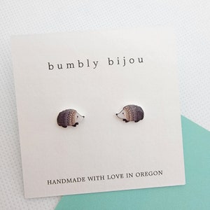 May include: A pair of hedgehog stud earrings on a white card with the text "bumbly bijou" and "HANDMADE WITH LOVE IN OREGON". The earrings feature a detailed design with gray, brown, and white colors. The background is a light blue and white.