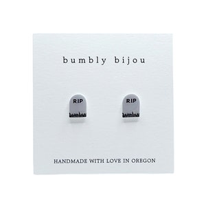 May include: A pair of white ceramic earrings shaped like tombstones with the text "RIP" on them. The earrings are on a white card with the text "bumbly bijou" and "HANDMADE WITH LOVE IN OREGON".