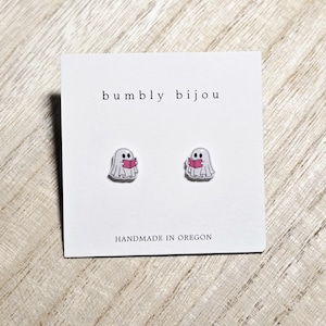 May include: A pair of ghost-shaped stud earrings, each holding a pink book. The earrings are on a white card with the text "bumbly bijou" and "HANDMADE IN OREGON". The earrings are a cute and whimsical accessory.