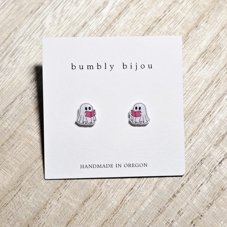 May include: A pair of ghost-shaped stud earrings, each holding a pink book. The earrings are on a white card with the text "bumbly bijou" and "HANDMADE IN OREGON". The earrings are a cute and whimsical accessory.