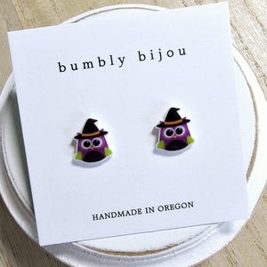 May include: A pair of purple owl earrings with black hats and green accents. The earrings are on a white card with the text "bumbly bijou" and "HANDMADE IN OREGON".