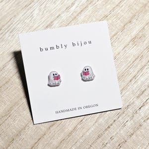 May include: A pair of ghost-shaped stud earrings. Each earring features a white ghost holding a pink book. The earrings are displayed on a white card with the text "bumbly bijou" and "HANDMADE IN OREGON".