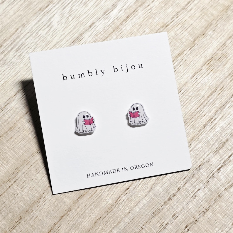 May include: A pair of ghost-shaped stud earrings. Each earring features a white ghost holding a pink book. The earrings are displayed on a white card with the text "bumbly bijou" and "HANDMADE IN OREGON".