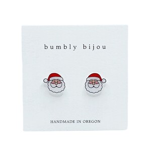 Puede incluir: Un par de pendientes de bot&oacute;n de Santa Claus blancos con gorros rojos y barbas blancas. Los pendientes est&aacute;n en una tarjeta blanca con el texto "bumbly bijou" y "HANDMADE IN OREGON".