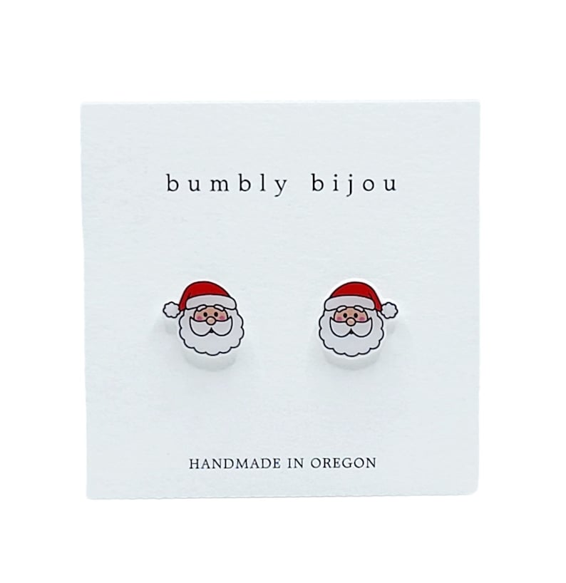 Puede incluir: Un par de pendientes de bot&oacute;n de Santa Claus blancos con gorros rojos y barbas blancas. Los pendientes est&aacute;n en una tarjeta blanca con el texto "bumbly bijou" y "HANDMADE IN OREGON".