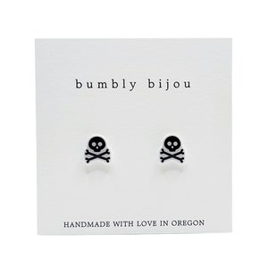May include: A pair of black skull and crossbones stud earrings on a white card with the text "bumbly bijou" and "HANDMADE WITH LOVE IN OREGON".