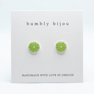 May include: A pair of lime slice stud earrings on a white card. The earrings are bright green with white details, resembling a cross-section of a lime. The card reads "bumbly bijou" and "HANDMADE WITH LOVE IN OREGON".