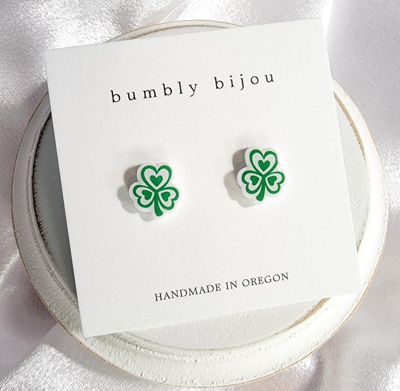 K&ouml;nnte beinhalten: Ein Paar gr&uuml;ne Kleeblatt-Ohrstecker auf einer wei&szlig;en Karte mit dem Text "bumbly bijou" und "HANDMADE IN OREGON".