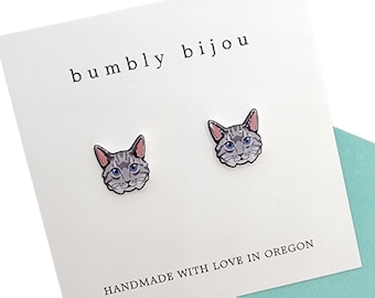 Może przedstawiać: Para srebrnych kolczyków z motywem szarej kociej twarzy. Kolczyki są prezentowane na białej kartce z tekstem "bumbly bijou" i "HANDMADE WITH LOVE IN OREGON".