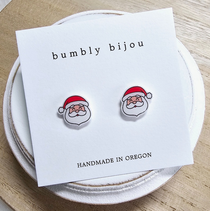 May include: A pair of white Santa Claus stud earrings with red hats and white beards. The earrings are on a white card with the text "bumbly bijou" and "HANDMADE IN OREGON".