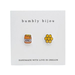 May include: A pair of honey-themed earrings with a honey jar and honeycomb design. The earrings are handmade in Oregon and feature the text "bumbly bijou" and "HANDMADE WITH LOVE IN OREGON".