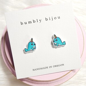 May include: A pair of blue narwhal stud earrings with a yellow and pink horn. The earrings are on a white card with the text "bumbly bijou" and "HANDMADE IN OREGON".