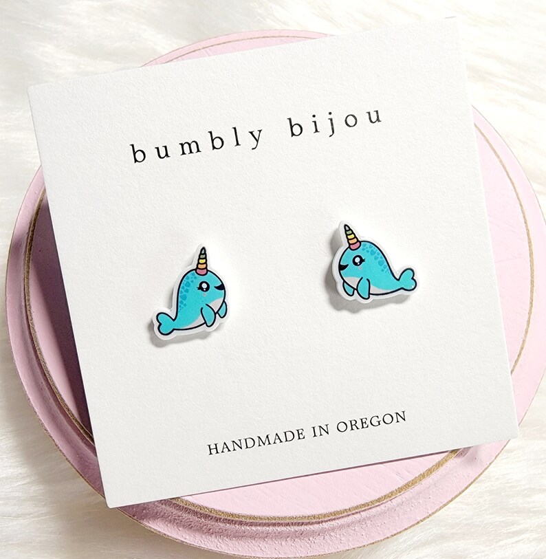 May include: A pair of blue narwhal stud earrings with a yellow and pink horn. The earrings are on a white card with the text "bumbly bijou" and "HANDMADE IN OREGON".