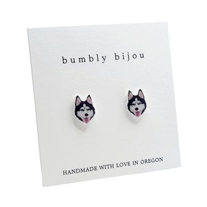 Op de afbeelding: Een paar zilveren oorbel studs met een zwart-witte husky hondenkop ontwerp. De oorbellen zijn op een witte kaart met de tekst "bumbly bijou" en "HANDMADE WITH LOVE IN OREGON".