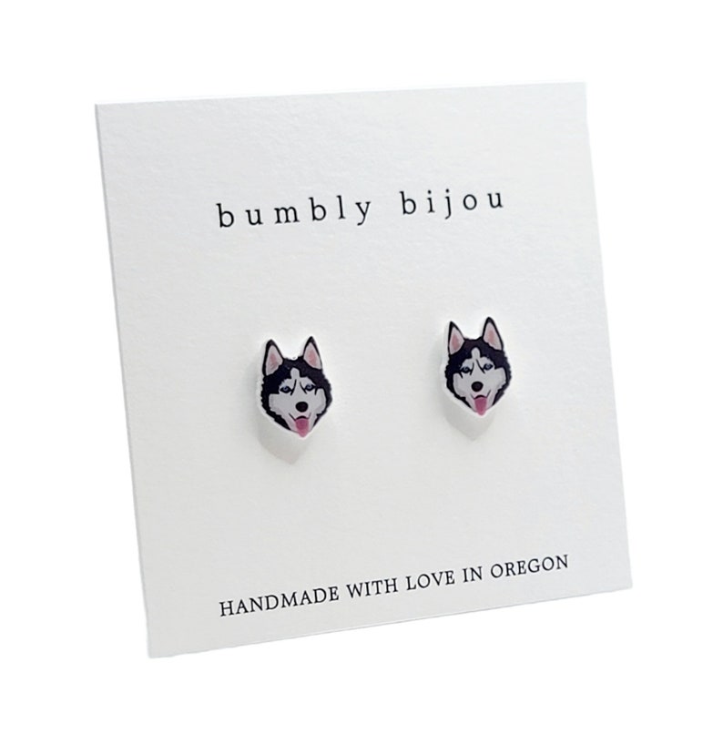 Op de afbeelding: Een paar zilveren oorbel studs met een zwart-witte husky hondenkop ontwerp. De oorbellen zijn op een witte kaart met de tekst "bumbly bijou" en "HANDMADE WITH LOVE IN OREGON".