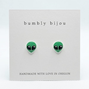 May include: A pair of green alien face stud earrings on a white card with the text "bumbly bijou" and "HANDMADE WITH LOVE IN OREGON".