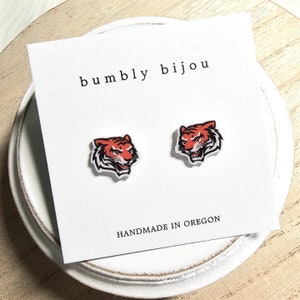 May include: A pair of tiger face stud earrings on a white card with the text "bumbly bijou" and "HANDMADE IN OREGON".