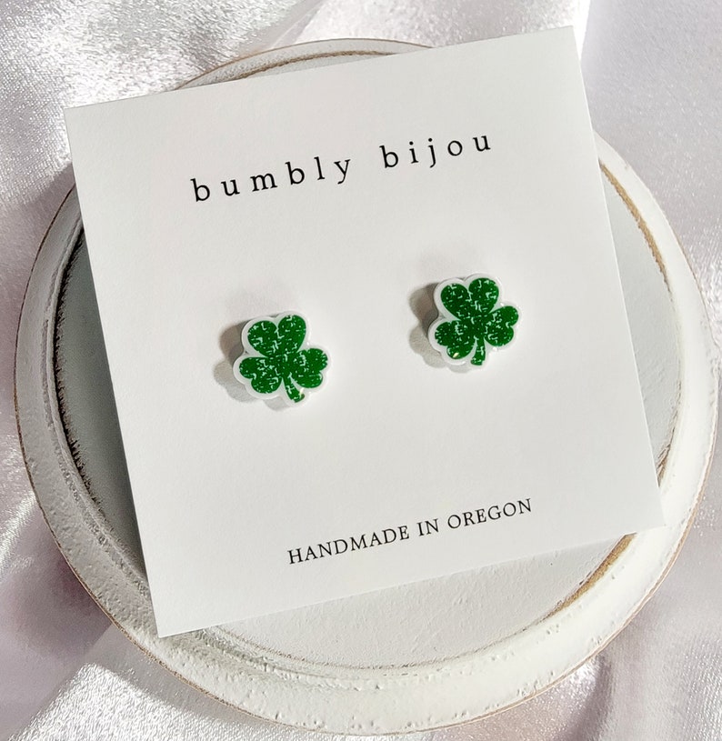 K&ouml;nnte beinhalten: Ein Paar gr&uuml;ne Glitzer-Kleeblatt-Ohrstecker auf einer wei&szlig;en Karte mit dem Text "bumbly bijou" und "HANDMADE IN OREGON".