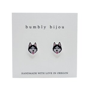 Op de afbeelding: Een paar zilveren oorbelletjes met een zwart-witte husky-hondengezichtsontwerp. De oorbelletjes bevinden zich op een witte kaart met de tekst "bumbly bijou" en "HANDMADE WITH LOVE IN OREGON".