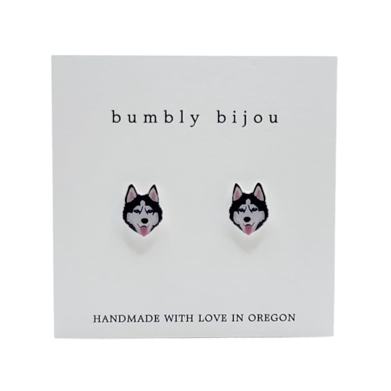 Op de afbeelding: Een paar zilveren oorbelletjes met een zwart-witte husky-hondengezichtsontwerp. De oorbelletjes bevinden zich op een witte kaart met de tekst "bumbly bijou" en "HANDMADE WITH LOVE IN OREGON".
