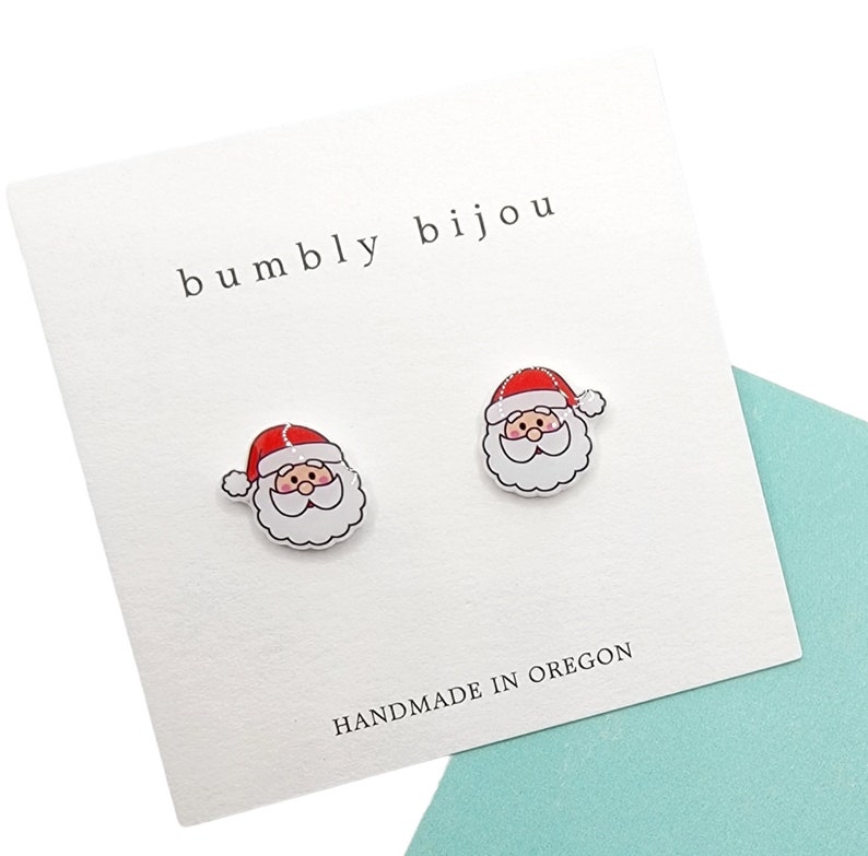 Puede incluir: Un par de pendientes de bot&oacute;n de Santa Claus blanco con gorros rojos y barbas blancas. Los pendientes est&aacute;n en una tarjeta blanca con el texto "bumbly bijou" y "HANDMADE IN OREGON".