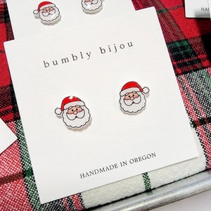 Puede incluir: Un par de pendientes blancos de Pap&aacute; Noel con gorros rojos y barbas blancas. Los pendientes est&aacute;n en una tarjeta blanca con el texto "bumbly bijou" y "HANDMADE IN OREGON".