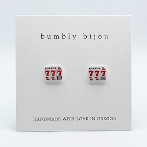 May include: A pair of white stud earrings with a red and white 777 slot machine design. The earrings are handmade in Oregon.
