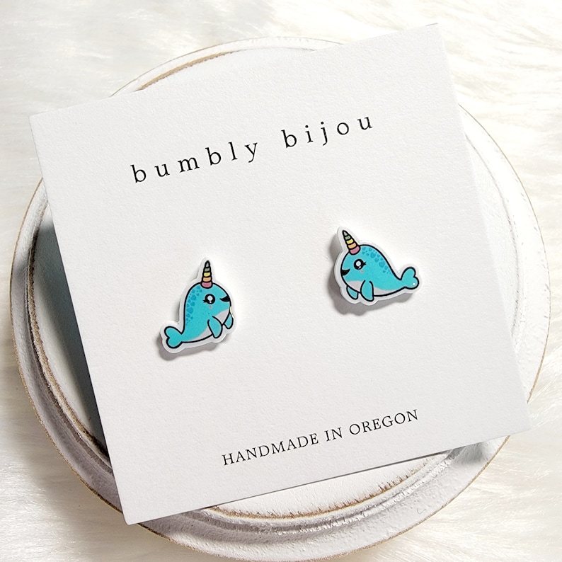 May include: A pair of blue and white narwhal stud earrings with a rainbow horn. The earrings are on a white card with the text "bumbly bijou" and "HANDMADE IN OREGON".
