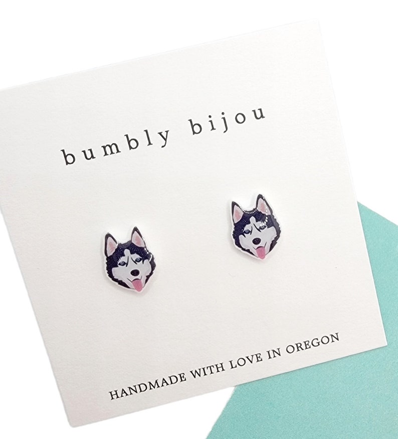 Op de afbeelding: Een paar zilveren oorbel studs met een zwart-witte husky hondenkop ontwerp. De oorbellen zijn verpakt op een witte kaart met de tekst "bumbly bijou" en "HANDMADE WITH LOVE IN OREGON".