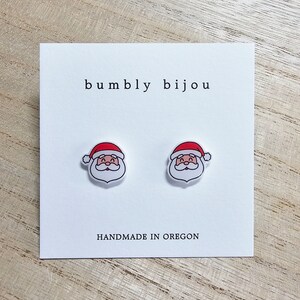 Peut inclure: Une paire de boucles d'oreilles blanches en forme de P&egrave;re No&euml;l avec des chapeaux rouges. Les boucles d'oreilles sont sur une carte blanche avec le texte "bumbly bijou" et "HANDMADE IN OREGON".