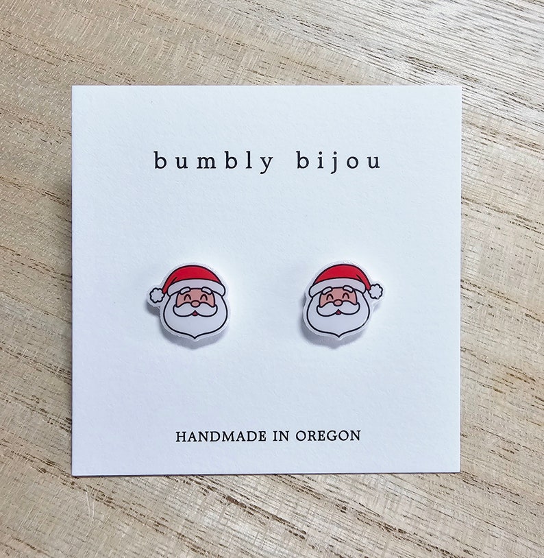 Peut inclure: Une paire de boucles d'oreilles blanches en forme de P&egrave;re No&euml;l avec des chapeaux rouges. Les boucles d'oreilles sont sur une carte blanche avec le texte "bumbly bijou" et "HANDMADE IN OREGON".