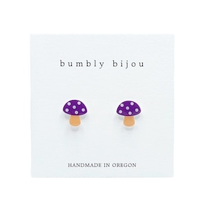 May include: A pair of purple mushroom stud earrings with white polka dots and orange stems. The earrings are handmade in Oregon and are displayed on a white card with the text "bumbly bijou" and "HANDMADE IN OREGON".
