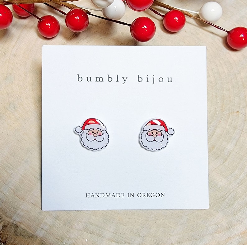 May include: A pair of white stud earrings with a cartoon image of Santa Claus's face. The earrings are on a white card with the text "bumbly bijou" and "HANDMADE IN OREGON".