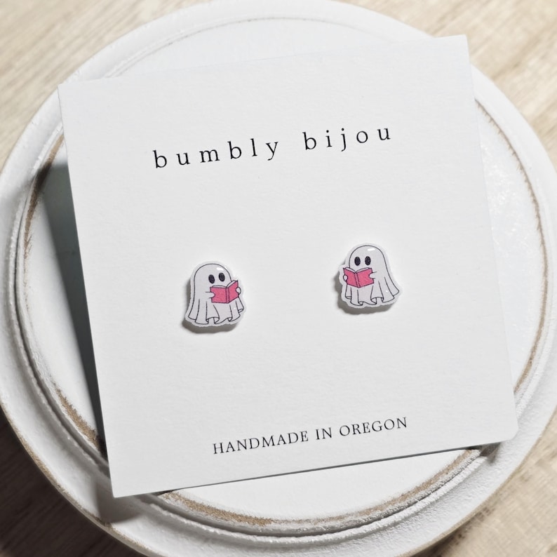 May include: A pair of ghost-shaped stud earrings. Each earring features a white ghost reading a pink book. The earrings are on a white card with the text "bumbly bijou" and "HANDMADE IN OREGON".