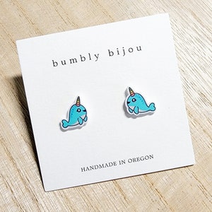 May include: A pair of narwhal stud earrings on a white card with the text "bumbly bijou" and "HANDMADE IN OREGON". The narwhals are blue with a yellow and orange horn.