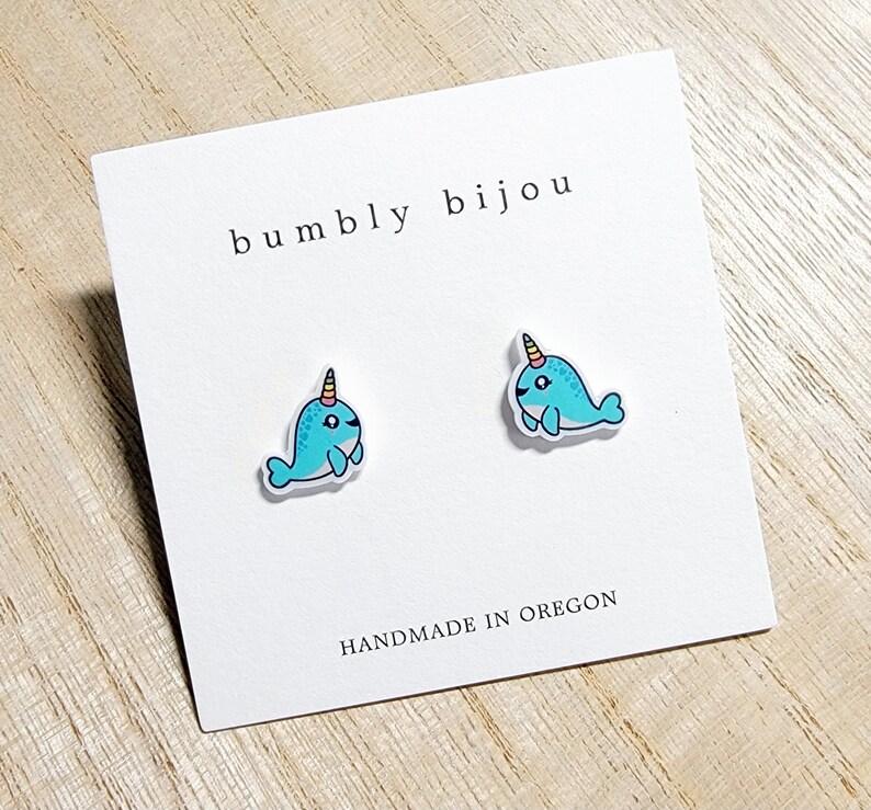 May include: A pair of narwhal stud earrings on a white card with the text "bumbly bijou" and "HANDMADE IN OREGON". The narwhals are blue with a yellow and orange horn.