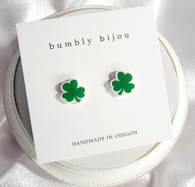 K&ouml;nnte beinhalten: Ein Paar gr&uuml;ne Kleeblatt-Ohrstecker auf einer wei&szlig;en Karte mit dem Text "bumbly bijou" und "HANDMADE IN OREGON".