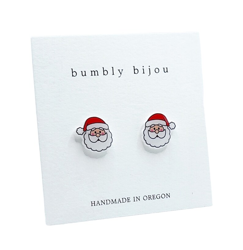 Puede incluir: Un par de pendientes de bot&oacute;n blancos con una cara de Pap&aacute; Noel de dibujos animados en cada uno. Los pendientes est&aacute;n en una tarjeta blanca con el texto "bumbly bijou" y "HANDMADE IN OREGON".