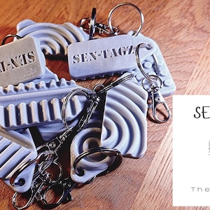 May include: A collection of grey and white keychains with different textures and designs. The keychains feature the text "SEN-TAGZ" and "SEN-TAGZ The Power of Touch".