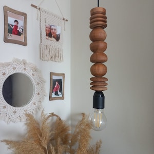 Puede incluir: Una l&aacute;mpara colgante de madera con una serie de elementos de madera redondeados apilados. La l&aacute;mpara cuelga de un cable negro y tiene una bombilla visible en la parte inferior. La madera tiene un tono c&aacute;lido y natural.