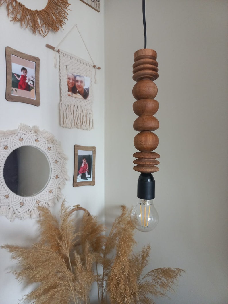 Puede incluir: Una l&aacute;mpara colgante de madera con una serie de elementos de madera redondeados apilados. La l&aacute;mpara cuelga de un cable negro y tiene una bombilla visible en la parte inferior. La madera tiene un tono c&aacute;lido y natural.