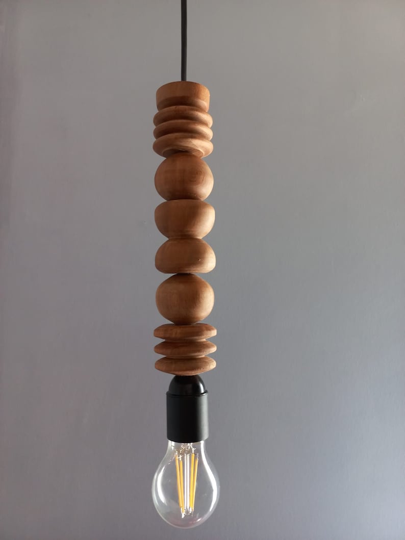 Puede incluir: Una l&aacute;mpara colgante de madera con una serie de elementos de madera torneada apilados en varias formas. La l&aacute;mpara tiene una base negra y una bombilla transparente con filamentos visibles. El cable es negro y el fondo es gris liso.