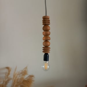 Puede incluir: L&aacute;mpara colgante de madera con dise&ntilde;o de cuentas apiladas. La l&aacute;mpara presenta una serie de cuentas de madera de diferentes formas. La bombilla es visible en la parte inferior y el cable es negro.