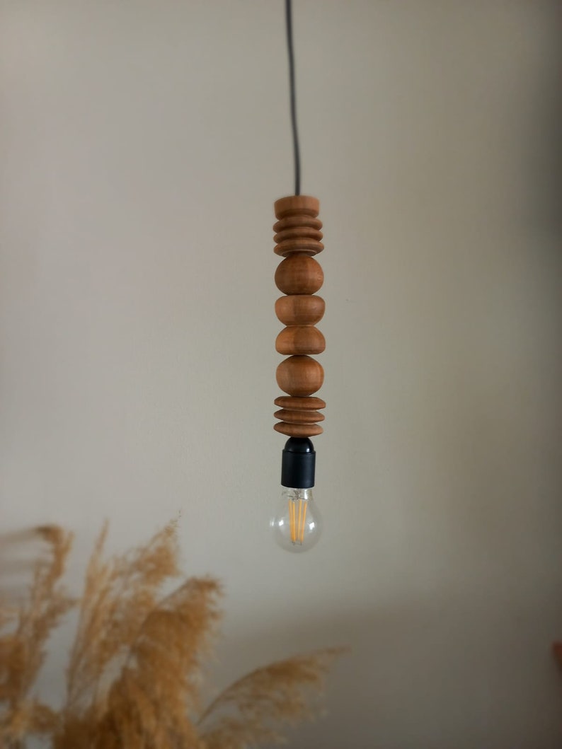 Puede incluir: L&aacute;mpara colgante de madera con dise&ntilde;o de cuentas apiladas. La l&aacute;mpara presenta una serie de cuentas de madera de diferentes formas. La bombilla es visible en la parte inferior y el cable es negro.