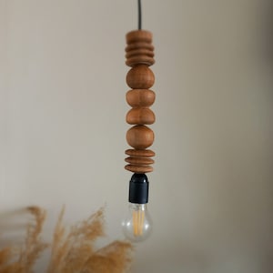 Puede incluir: L&aacute;mpara colgante de madera con una serie de cuentas de madera redondeadas y formas cil&iacute;ndricas apiladas. Un cable negro suspende la l&aacute;mpara, con un casquillo negro que sostiene una bombilla transparente. La luz est&aacute; suspendida sobre un fondo neutro.