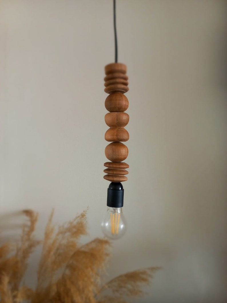 Puede incluir: L&aacute;mpara colgante de madera con una serie de cuentas de madera redondeadas y formas cil&iacute;ndricas apiladas. Un cable negro suspende la l&aacute;mpara, con un casquillo negro que sostiene una bombilla transparente. La luz est&aacute; suspendida sobre un fondo neutro.
