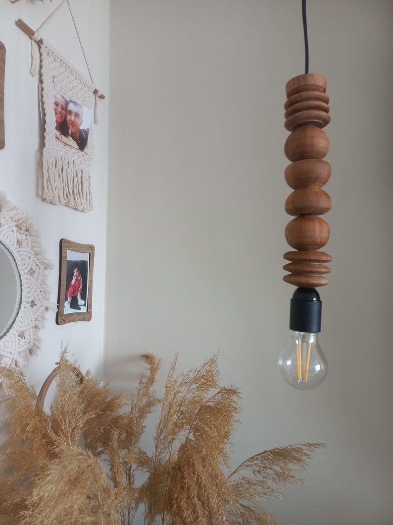 Puede incluir: Una l&aacute;mpara colgante de madera con un dise&ntilde;o cil&iacute;ndrico apilado. La l&aacute;mpara est&aacute; hecha de m&uacute;ltiples piezas de madera de varias formas y tama&ntilde;os, suspendidas de un cable negro. Una bombilla transparente es visible en la parte inferior.