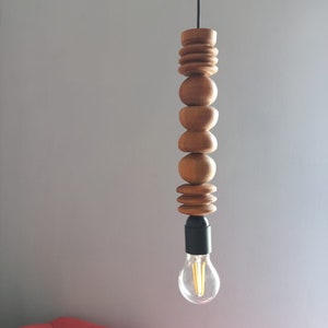 Puede incluir: L&aacute;mpara colgante de madera con una serie de elementos de madera redondeados apilados. La bombilla es visible en la parte inferior, con un accesorio negro. La luz cuelga de un cable negro sobre un fondo gris.