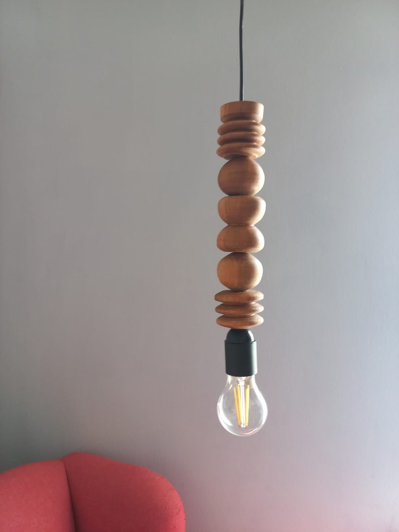 Puede incluir: L&aacute;mpara colgante de madera con una serie de elementos de madera redondeados apilados. La bombilla es visible en la parte inferior, con un accesorio negro. La luz cuelga de un cable negro sobre un fondo gris.